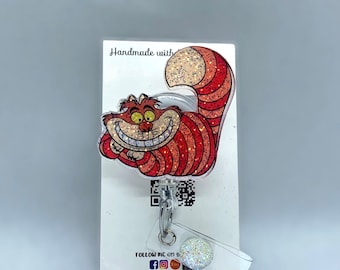 Op de afbeelding: Een rode en witte glitterende Cheshire Cat uit Alice in Wonderland is afgebeeld op een witte kaart met de tekst "Handmade with" en "Follow me on" met social media-iconen. De kaart is bevestigd aan een transparante, intrekbare badgehouder van plastic met een zilveren clip en een transparante, ronde hanger van plastic met glitter.