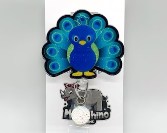 May include: A blue and green peacock with glitter accents on a white background. The peacock is attached to a clear retractable badge reel with a white glitter charm that says "Mooshino Designs".