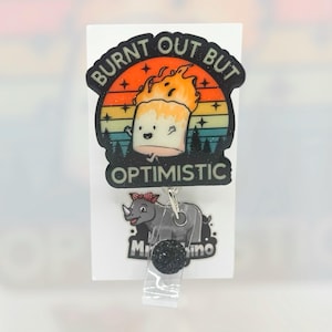 May include: A badge reel with a cartoon marshmallow on fire, with the text "BURNT OUT BUT OPTIMISTIC". Below is a rhino with a red bow and the text "Mr. Rhino". The reel has a black glitter button and a clear plastic strap.