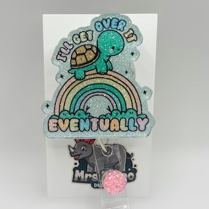 May include: A badge reel with a glittery light blue background, featuring a turtle on a rainbow with the text "I'll get over it eventually". The reel has a pink glitter button and a rhino graphic.
