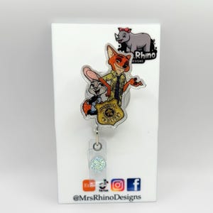 May include: A clear acrylic badge reel featuring the characters Judy Hopps and Nick Wilde from Zootopia. The badge reel has a glittery accent and a police badge design. The Rhino Designs logo is visible at the top, and social media icons are at the bottom.