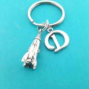 May include: Silver keychain with a rocket ship charm and a letter D charm. The keychain is on a turquoise background.