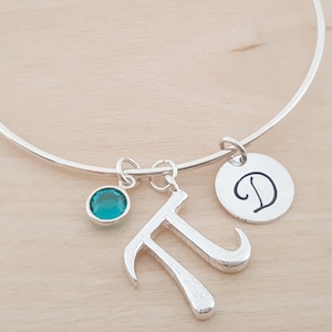 May include: Silver adjustable bangle bracelet with a teal-colored charm, a letter "T" charm, and a circular charm with the letter "D". The bracelet is displayed on a light wood surface.