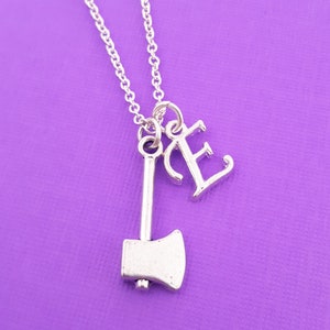 May include: A silver necklace with a small axe charm and a letter 'E' charm. The necklace is on a purple background.