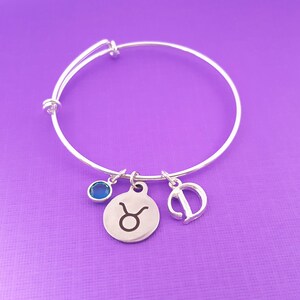 May include: Silver adjustable bangle bracelet with three charms. The charms include a blue gemstone, a circular charm with the Taurus zodiac symbol, and a charm with the letter "T". The bracelet is displayed on a purple background.