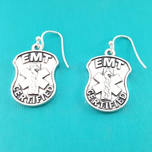May include: Silver earrings with a silver shield-shaped charm that says "EMT CERTIFIED" with a medical symbol in the center.