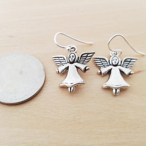 May include: A pair of silver-colored angel earrings. Each earring features a small angel figure with outstretched wings and a halo. The earrings are shown on a light wood surface next to a quarter for size comparison.
