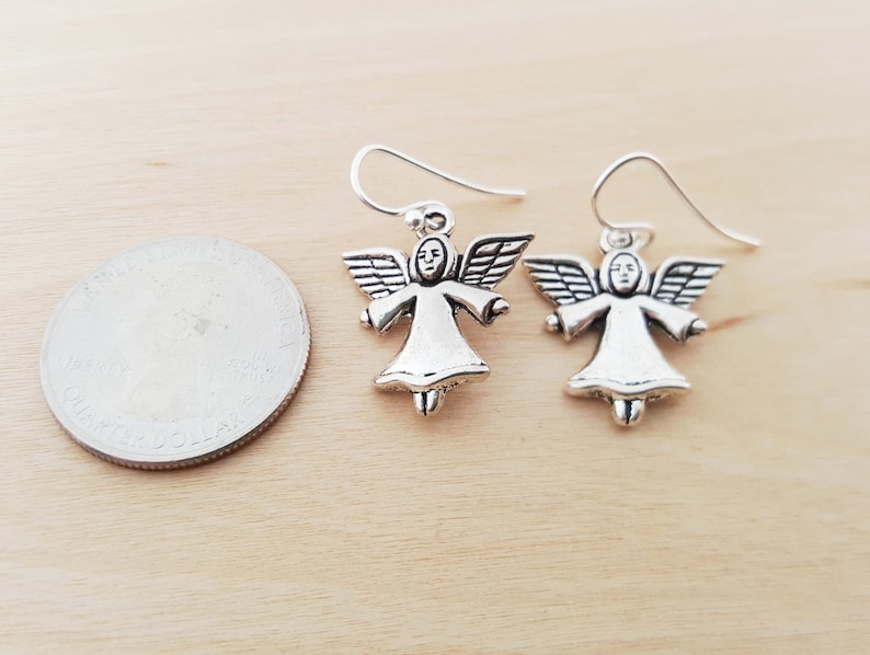 May include: A pair of silver-colored angel earrings. Each earring features a small angel figure with outstretched wings and a halo. The earrings are shown on a light wood surface next to a quarter for size comparison.