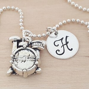 May include: Silver necklace with a charm of a drum set and a round charm with the letter 'H' engraved on it.