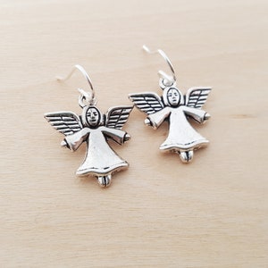 May include: A pair of silver-colored angel earrings. Each earring features a small angel figure with outstretched wings, a simple face, and a flowing gown. The earrings hang from silver-toned ear wires, set against a light wood grain background.