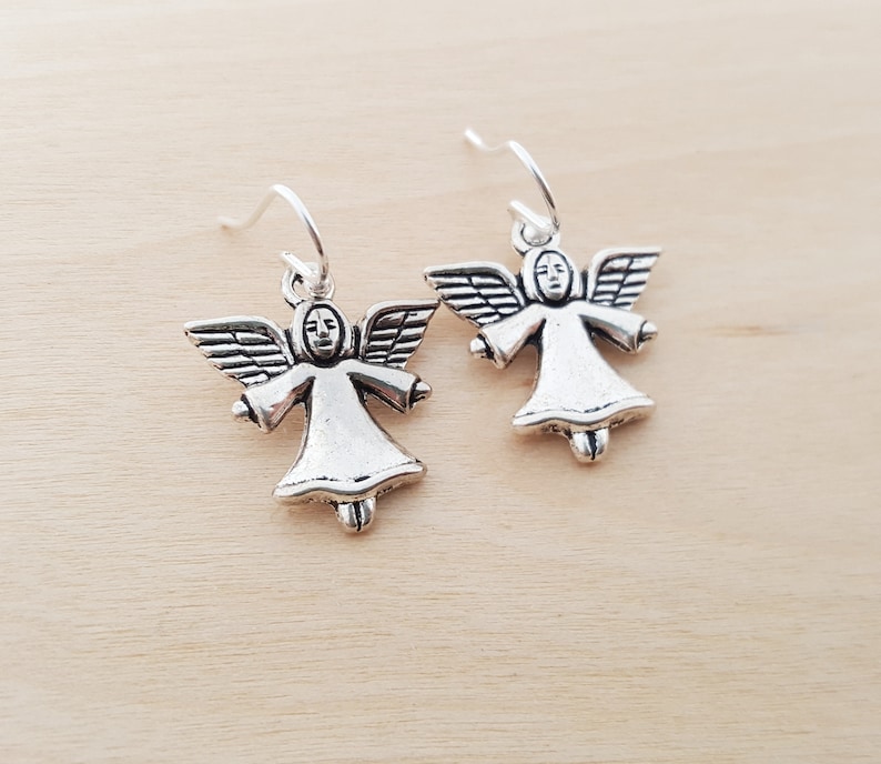 May include: A pair of silver-colored angel earrings. Each earring features a small angel figure with outstretched wings, a simple face, and a flowing gown. The earrings hang from silver-toned ear wires, set against a light wood grain background.
