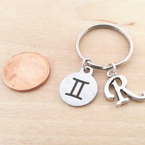 May include: Silver keychain with a Gemini zodiac sign charm and a letter R charm. The charms are attached to a silver key ring.