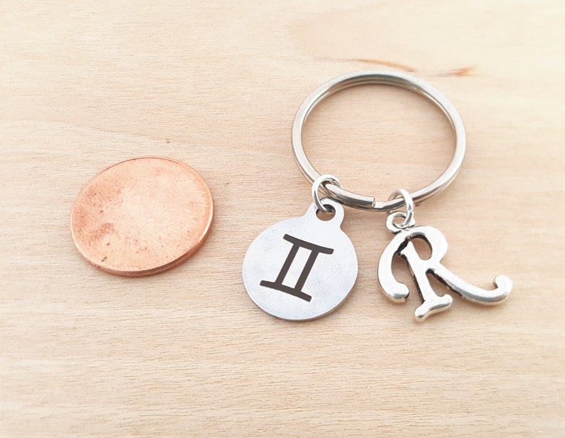 May include: Silver keychain with a Gemini zodiac sign charm and a letter R charm. The charms are attached to a silver key ring.