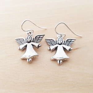 May include: A pair of silver-colored angel earrings. Each earring features a small angel figure with outstretched arms and detailed wings. The earrings have a simple hook design for pierced ears, set against a light wooden background.