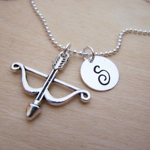 May include: Silver necklace with a bow and arrow charm and a round charm with the letter 'S' engraved on it.