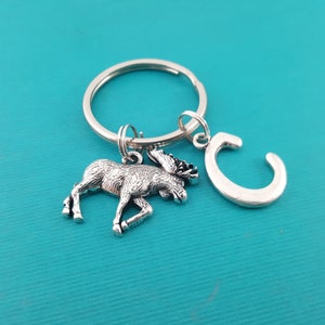 May include: Silver moose keychain with a silver letter C charm attached to a silver key ring. The keychain is on a turquoise background.
