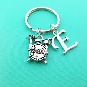 May include: Silver keychain with a drum set charm and the letter "E" hanging from a silver key ring.