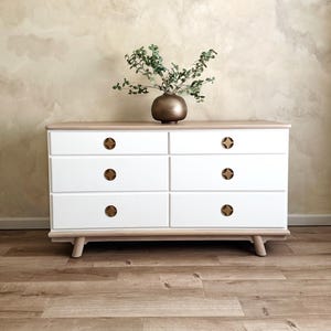 May include: White dresser with six drawers and gold-toned knobs. The dresser has a light wood top and sits on four legs. A vase with green leaves sits on top of the dresser.