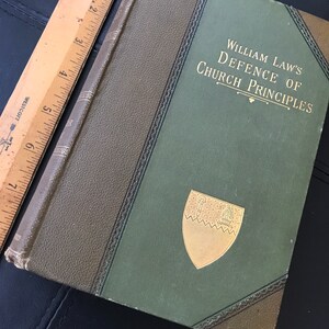 Puede incluir: Un libro de tapa dura verde con letras doradas y un escudo dorado en la portada. El título del libro es "William Law's Defence of Church Principles".