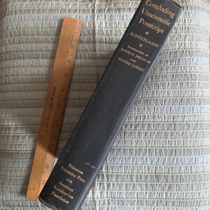 May include: A vintage book titled "Concluding Unscientific Postscript" by Kierkegaard, translated by David F. Swenson and Walter Lowrie, published by Princeton University Press. A wooden ruler, approximately 12 inches long, rests beside the book.