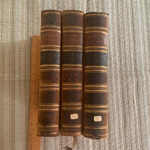 Op de afbeelding: Drie antieke boeken met bruinleren covers en gouden letters. De ruggen lezen "HOOKER'S WORKS" en "VOL. I, II, III". De boeken hebben decoratieve gouden banden en zijn naast elkaar geplaatst. Een houten liniaal is links zichtbaar.