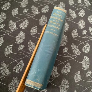 以下が含まれることがあります： ギネス著「The Divine Programme of the World's History」というタイトルのヴィンテージ本。本は、金色の文字と装飾的なボーダーが付いた水色の表紙です。白い花柄の暗い背景に、木製の定規で支えられています。