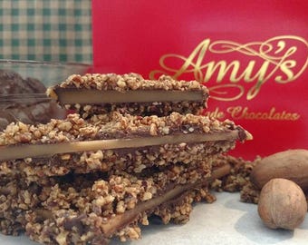May include: Close-up of toffee candy stacked on top of each other, covered in chopped nuts. A red box with gold lettering that reads "Amy's Handmade Chocolates" is in the background, along with several whole pecans.