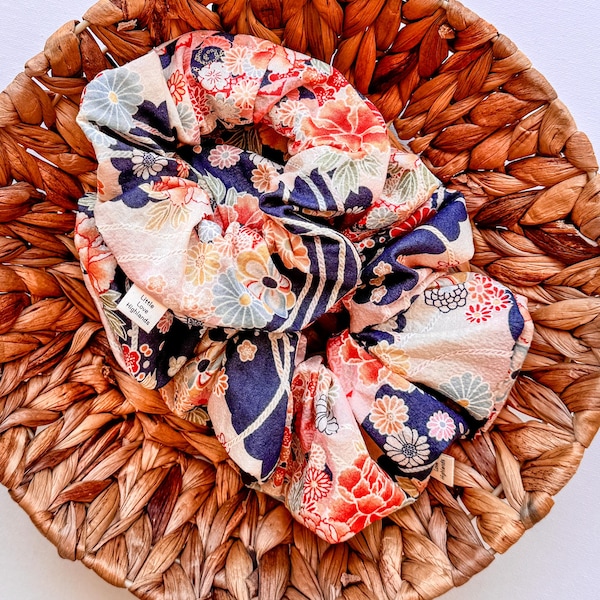 May include: A navy blue and cream floral patterned hair scrunchie with a white tag that reads "Little Luxe Highlands".