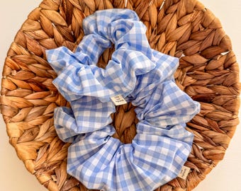 May include: Two blue and white gingham scrunchies are displayed on a woven wicker basket. The scrunchies have a light blue and white checkered pattern. Each scrunchie has a small tag that reads "Little Low Highlands".