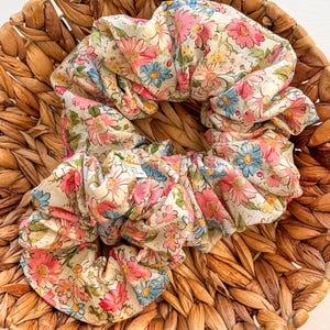 May include: Two floral scrunchies with pink, blue, and yellow flowers on a cream background. The scrunchies are in a woven basket. The scrunchies are made of a soft, lightweight fabric.