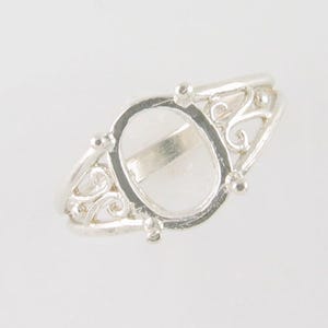 May include: A silver ring with an oval setting and ornate filigree detailing. The ring features a clear, oval-shaped space in the center, and small decorative balls are placed around the setting. The band has a delicate, openwork design.