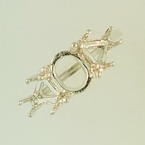 May include: A silver-colored ring setting with a central circular design and multiple prongs. The setting is designed to hold a gemstone. The ring setting is made of a shiny metal and has a detailed, ornate design.