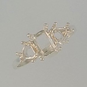 May include: A silver ring setting with a central square and two side settings, one triangular and one round. The ring has small prongs for holding gemstones. The ring is shown against a light gray background.