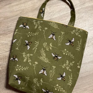 May include: A green canvas tote bag with a bee and floral pattern. The bag has a yellow top stitch and two handles.