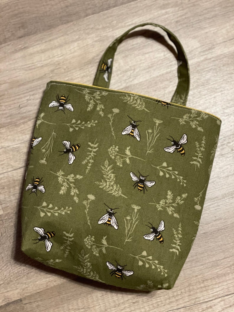 May include: A green canvas tote bag with a bee and floral pattern. The bag has a yellow top stitch and two handles.