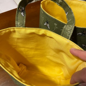 May include: Two green and yellow fabric storage bins with bee and flower designs. The bins have handles and are lined with yellow fabric.