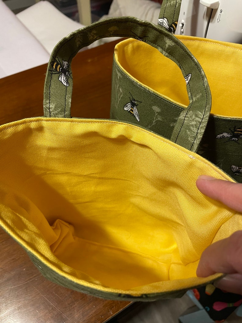 May include: Two green and yellow fabric storage bins with bee and flower designs. The bins have handles and are lined with yellow fabric.