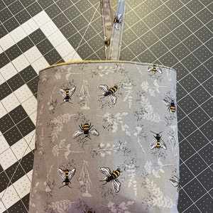 May include: A gray fabric hanging storage bin with a yellow trim and a gray fabric handle. The bin is decorated with a repeating pattern of bees and flowers.