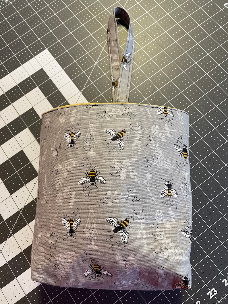 May include: A gray fabric hanging storage bin with a yellow trim and a gray fabric handle. The bin is decorated with a repeating pattern of bees and flowers.