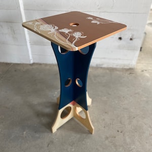 May include: A small, square, wooden table with a blue and beige base. The top of the table is painted a light brown color and features a white floral design.