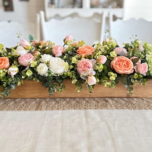 May include: A wooden box filled with a floral arrangement of pink, peach, and white roses, and greenery. The box is sitting on a table with a burlap runner.