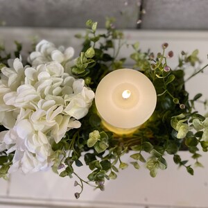 Puede incluir: Un centro de mesa con una vela blanca, una hortensia blanca y vegetaci&oacute;n verde de eucalipto. La vela est&aacute; encendida y el centro de mesa est&aacute; en una caja de madera.