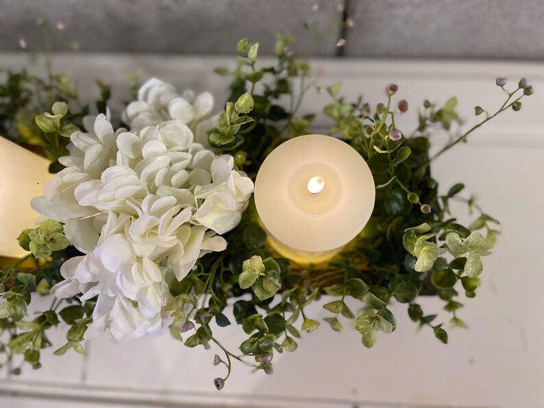 Puede incluir: Un centro de mesa con una vela blanca, una hortensia blanca y vegetaci&oacute;n verde de eucalipto. La vela est&aacute; encendida y el centro de mesa est&aacute; en una caja de madera.