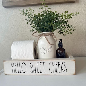 May include: White wooden box with the text "Hello Sweet Cheeks" on the front. The box contains a roll of toilet paper, a clear glass jar with a white lid and the word "Ball" on it, and a small brown glass bottle with a spray nozzle.