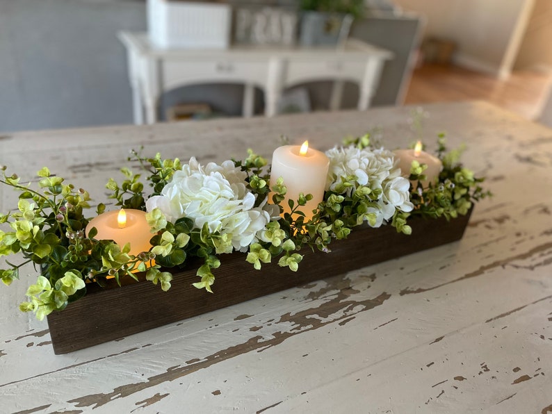 Puede incluir: Un centro de mesa de madera con hortensias blancas y vegetaci&oacute;n, decorado con tres velas blancas.