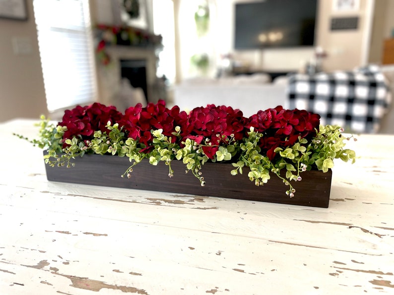 May include: A dark brown wooden box planter filled with red hydrangea flowers and green foliage. The planter is sitting on a white distressed table.