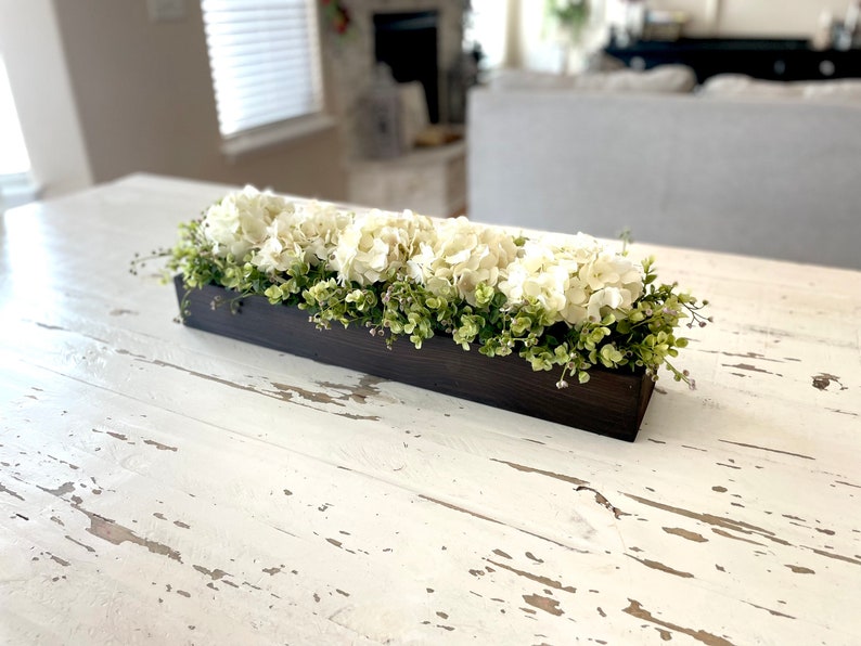 May include: A wooden box planter filled with white hydrangea flowers and green foliage. The planter is sitting on a white distressed table.