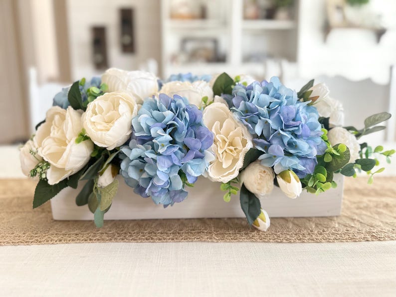 K&ouml;nnte beinhalten: Eine rechteckige, wei&szlig;e Holzkiste gef&uuml;llt mit k&uuml;nstlichen Blumen. Das Arrangement zeigt blaue Hortensien, cremefarbene Pfingstrosen und wei&szlig;e Rosen, akzentuiert mit gr&uuml;nen Bl&auml;ttern. Die Kiste steht auf einem Jutel&auml;ufer.