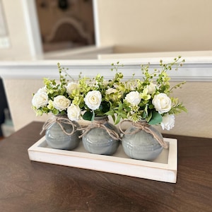 May include: Three white faux flower arrangements in gray ceramic vases with burlap accents, displayed on a white wooden tray. The tray is on a dark brown wooden surface.