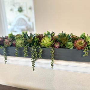 May include: A rectangular, dark gray planter box filled with a variety of artificial succulents. The succulents are in shades of green, burgundy, and pink, with trailing greenery. A decorative piece for home decor.